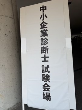A white sign with black Japanese text reading "中小企業診断士 試験会場" (Small and Medium Enterprise Consultant Examination Venue) stands outside a building, indicating the location of an entrance exam.