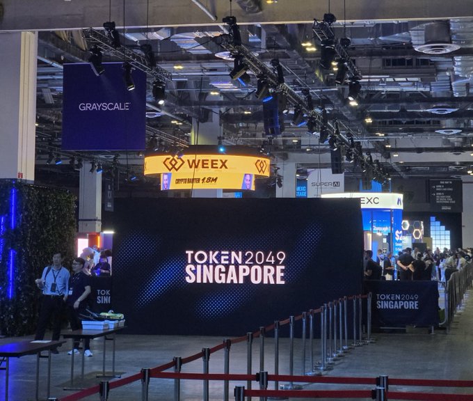 A convention hall entrance featuring a large 'TOKEN2049 SINGAPORE' display, with banners for 'GRAYSCALE' and 'WEEX 1.8M' visible above the crowd.