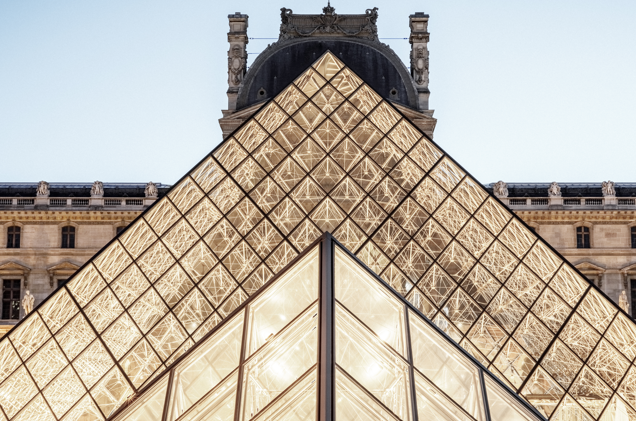An upward view of the Louvre Pyramid at night, with its illuminated glass panels contrasting against the historic palace facade.