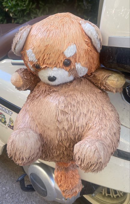 A worn, brown plush dog toy with white patches around its eyes sits on the seat of a motorcycle or scooter.