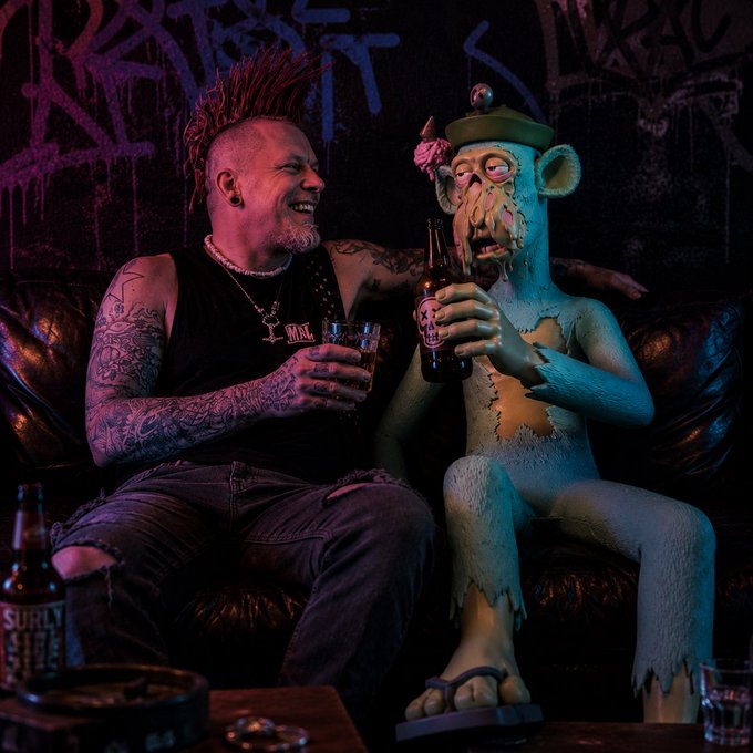 A punk-styled man with a red mohawk sits on a leather couch laughing alongside a grotesque green ape statue wearing a beret, sharing beers. A beer bottle labeled "SURLY" sits in the foreground against a graffiti-covered wall, matching the edgy "Mutant Monday" punk vibe of the post.