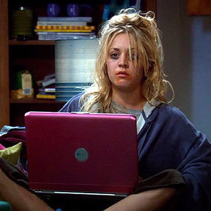 A young woman with messy blonde hair looking at a pink laptop with a surprised expression, sitting in a room with shelves in the background.