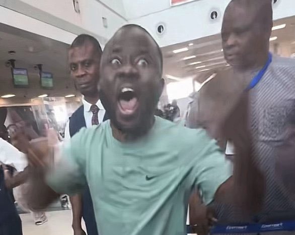 A photograph of a young man in a light green Nike t-shirt making an exaggerated shocked face with wide eyes and open mouth, with two other men visible behind him in what appears to be a public indoor space.
