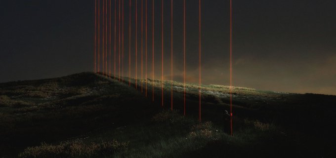 Red laser beams shoot down onto a dark, grassy hillside at night, with a small silhouetted figure standing among the vertical light columns in this installation by Outrun Youth titled "Hidden Geometry."