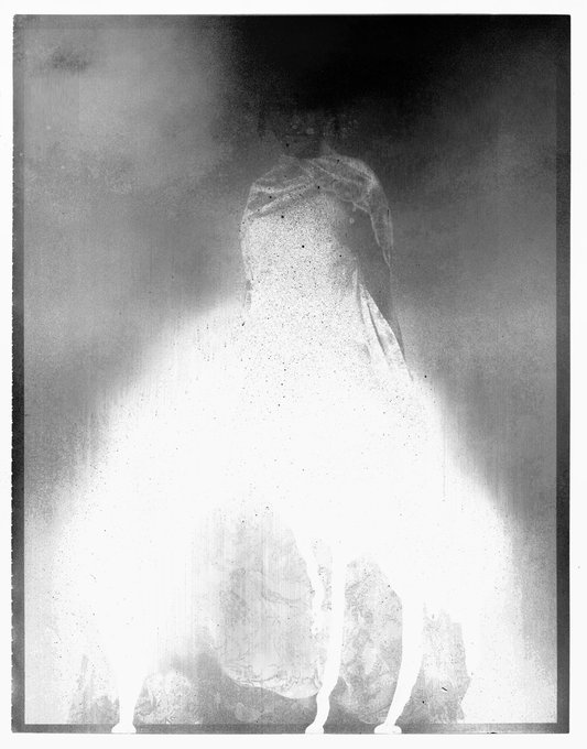A grainy, high-contrast black-and-white photograph showing a ghostly, faded figure standing behind a white curtain, with the image having a vintage, overexposed aesthetic.