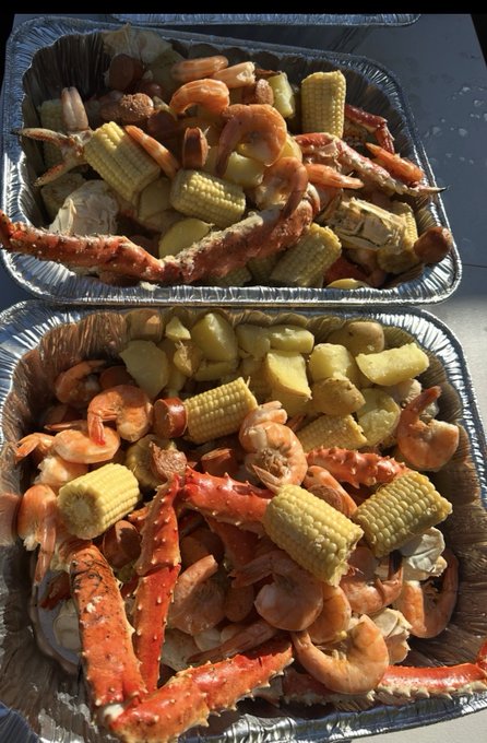 Two foil trays filled with a seafood boil containing crab legs, shrimp, corn on the cob, and potatoes.