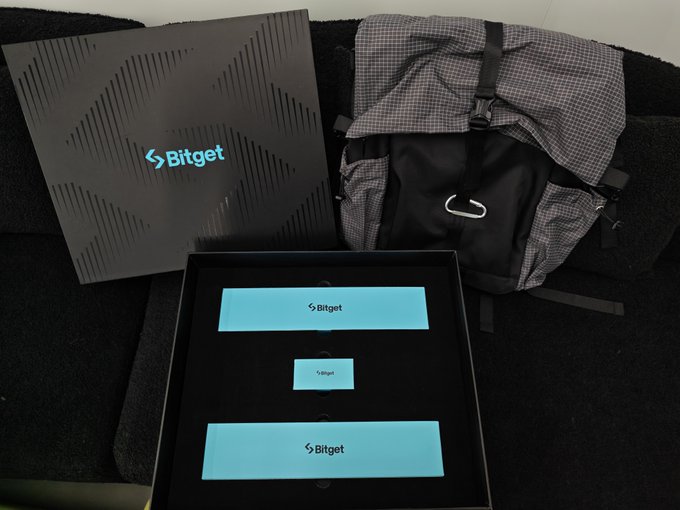 A 'Bitget' branded gift box with branded cards next to a gray Nike backpack on a dark surface.