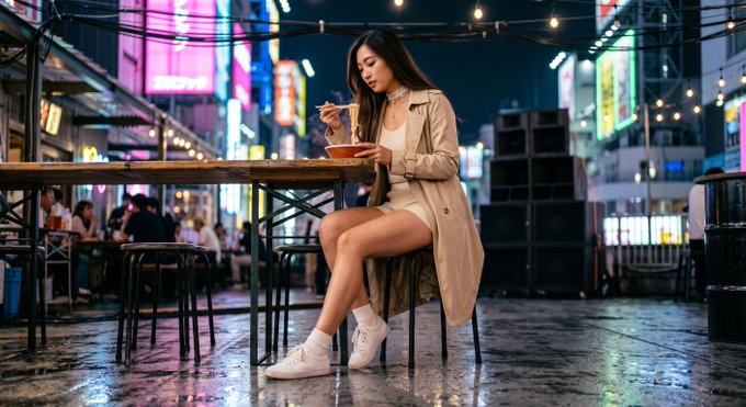 A young woman sits at an outdoor table eating from a bowl with chopsticks in a neon-lit city street at night, wearing a beige trench coat and white sneakers.