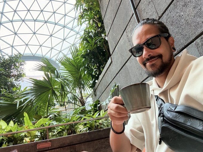 A man with sunglasses and a beard smiles while holding a metal mug inside a futuristic glass dome surrounded by lush tropical greenery, likely at Jewel Changi Airport. The setting fits the post's "GM Singapore" greeting as a stylish pre-event moment before Token2049.