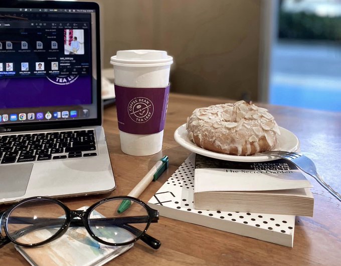 The image shows a wooden table with various items placed on top of it. There are two cups - one containing coffee and another holding tea. A doughnut is also present on the table. Additionally, there are three books arranged next to each other, along with a pair of glasses. The scene appears to be set in an office or workspace environment where someone might enjoy their breakfast while working or studying.