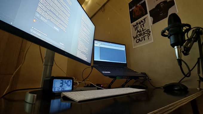 A dimly lit home office setup features a monitor displaying text, a laptop on a stand, a microphone, a keyboard, a mouse, and a digital clock showing 3:40, with posters on the wall reading 'WHAT IS IT ALL WORKS OUT?' and 'NO ONE IS STORIES'.