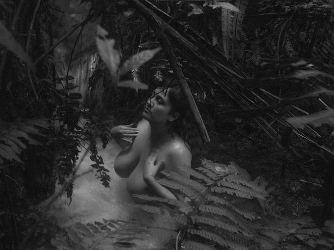 A nude woman partially draped in lush ferns gazes upward in a misty black-and-white jungle scene, evoking the ethereal, dreamlike quality suggested by the accompanying post about an NFT on objkt.com.