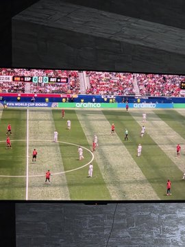 A TV screen showing a soccer match in progress on a green field, with advertising boards displaying 'aramco' and 'THE HOME OF FOOTBALL'.