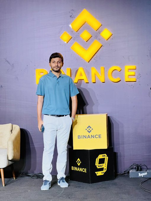 A man in a teal polo shirt and white pants poses in front of a Binance-branded backdrop with the company's yellow logo and promotional boxes.