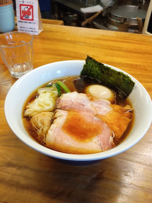 A bowl of ramen with chashu pork, wontons, a soft-boiled egg, nori, and vegetables in a rich broth, served at a wooden counter.