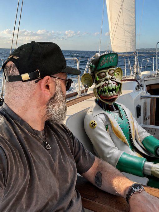 A bearded man wearing a black cap and sunglasses sits on a boat next to a large grotesque figure wearing a 'BAYC' cap and sunglasses.