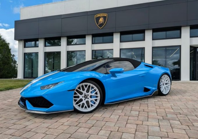A bright blue Lamborghini Huracán Spyder is parked in front of a Lamborghini dealership, its sleek convertible design and aggressive styling making it impossible to ignore—much like the song lyrics "I tried to walk away could you?" evoke the difficulty of leaving something so captivating behind.