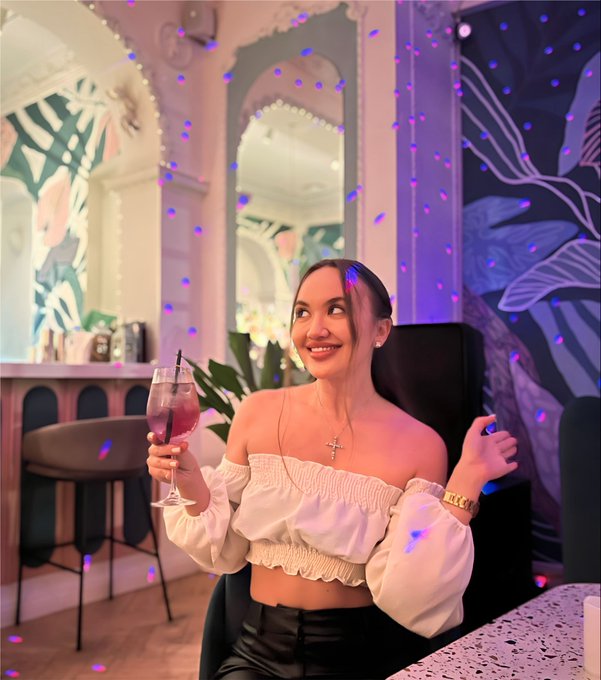 A smiling woman with dark hair wearing a white off-shoulder top and black leather pants sits holding a glass of red drink in a stylish cafe or bar setting with mirrors and purple ambient lighting.