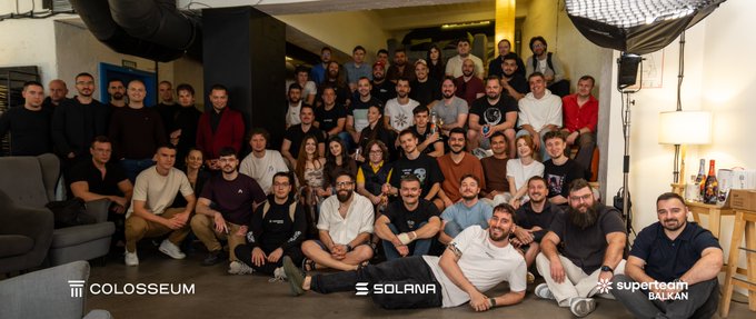 The image shows a large group of participants posing together at what appears to be the Colosseum hackathon, with logos for "COLOSSEUM," "SOLANA," and "Superteam BALKAN" displayed at the bottom. The diverse crowd, gathered in an indoor venue with photography equipment visible, reflects the community-focused event mentioned in the post about building an educational platform on Solana.