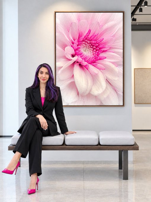 A woman with purple-streaked hair sits on a bench wearing a black suit with a pink top and bright pink heels, positioned in front of a large framed photograph of a pink flower.