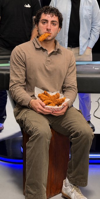 A man in a tan polo shirt sits on a stool eating fried chicken from a paper basket.