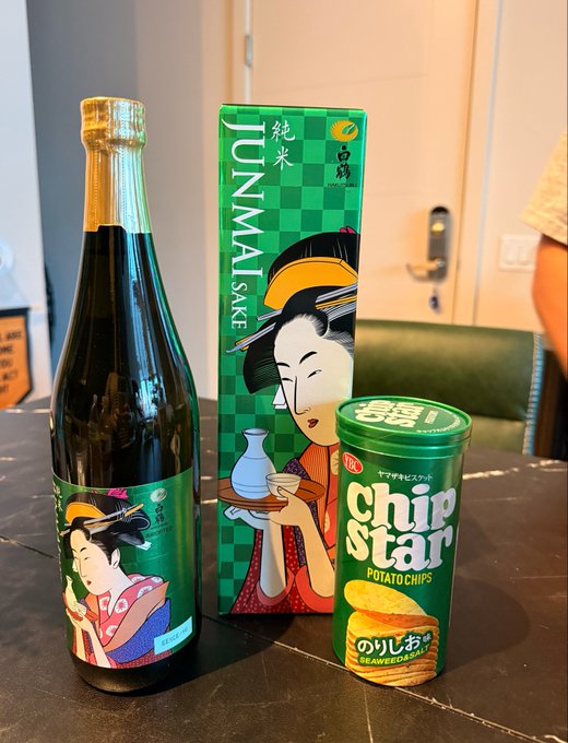 A bottle of Junmai sake and its green checkered box featuring a geisha illustration sit on a table next to a green canister of "Chip Star Potato Chips" in seaweed flavor (のりしお).