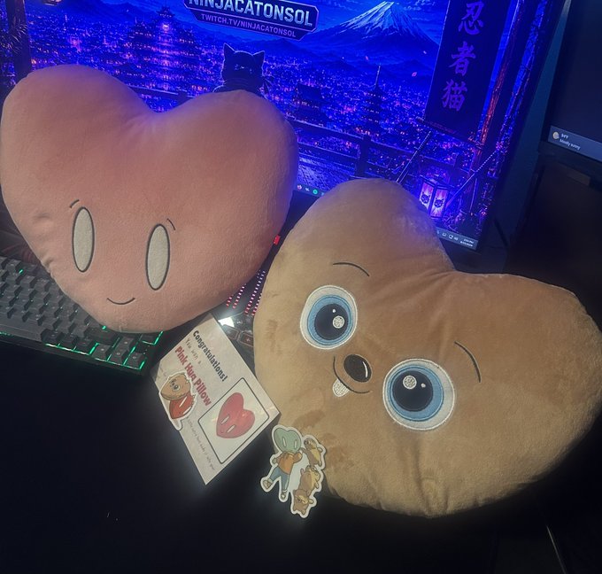 Two heart-shaped plush pillows—one pink with a simple smiley face and one brown with large blue eyes—sitting on a desk in front of a monitor displaying a Tokyo nighttime scene with Japanese text.