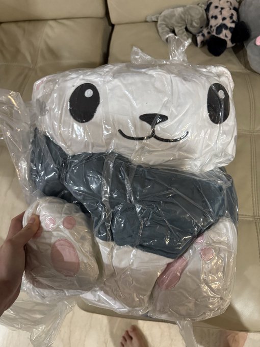A 1.5-metre Hypurr plush toy, still wrapped in clear plastic packaging, sits on a leather couch. The caption explains the owner is contemplating unwrapping the oversized stuffed animal, which was produced for the HypurrCo event at Token2049 and suggests 50 of them may appear at next year's gathering.