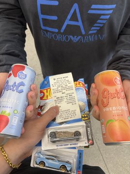 A person in an Emporio Armani EA7 shirt holding two beverage cans (Guava and Lychee flavors), small toy cars, and a receipt at what appears to be a store or shop.
