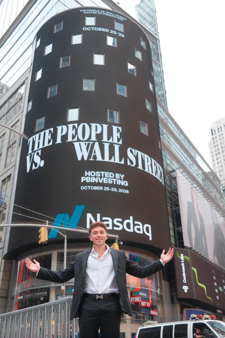 A man in a suit stands with open arms in front of the Nasdaq building in Times Square, where a large screen advertises 'The People vs. Wall Street' event hosted by PBINVESTING on October 25-29, 2025.