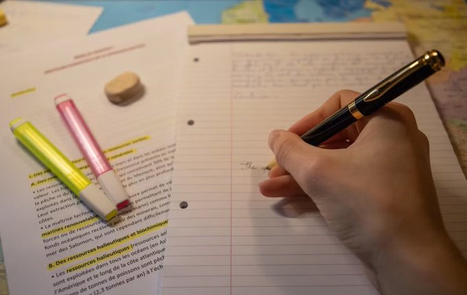 A close-up of a hand writing with a black fountain pen on lined notebook paper, with a French textbook, yellow and pink highlighters, and an eraser visible on the desk.