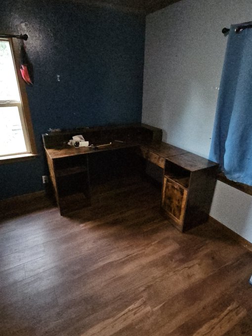A dimly lit room features a dark blue accent wall and a brown curtain by the window, with a large L-shaped wooden desk in the corner.