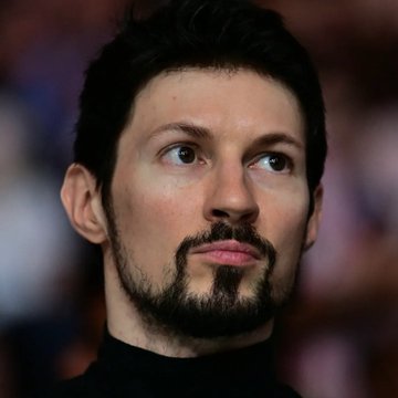 A close-up portrait of a man with short dark hair, a goatee, and brown eyes looking off to the side while wearing a black turtleneck.