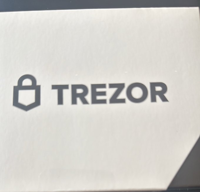 A close-up of a paper sleeve with the 'TREZOR' hardware wallet logo and padlock icon printed on it.