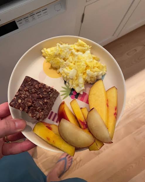 A first-person view of a hand holding a white plate containing scrambled eggs, a square of dark seeded crispbread, and several slices of fresh peach.