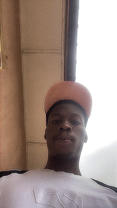 A low-angle selfie of a young man wearing a pink backwards baseball cap and a white t-shirt, standing near a building with a window frame visible behind him.