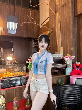 A young woman in an Argentina-style blue and white soccer jersey and white shorts stands in a retro-styled room with vintage electronics and decor.