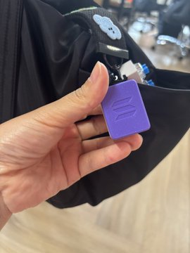 A hand holds a purple, block-shaped accessory clipped onto the strap of a black bag, featuring an embossed logo and a small cartoon keychain.