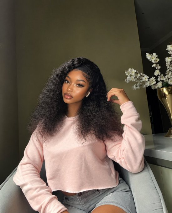 A young woman with long curly black hair sitting on a gray chair, wearing a pink cropped sweatshirt and denim shorts, posing against an olive green wall with a vase of white flowers on a table beside her.