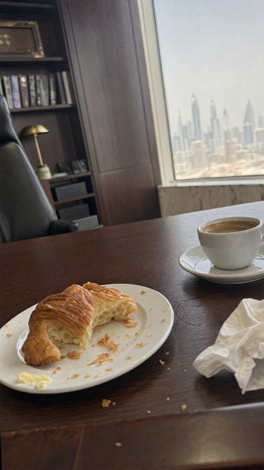 A partially eaten croissant with butter on a white plate beside a cup of coffee, set on a dark wooden table with a city skyline view through a large window in the background.