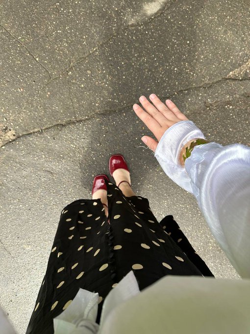 A first-person photo looking down at red Mary Jane shoes, a black polka dot skirt, and a white-sleeved arm extended outward on a paved surface.
