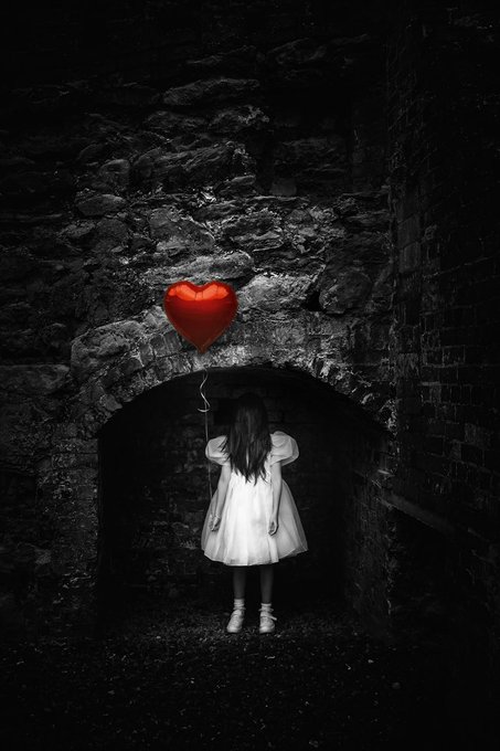 A young girl in a white dress stands with her back turned, holding a vivid red heart-shaped balloon that floats above her in a dark, shadowy stone archway.