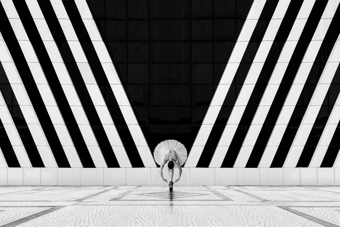 A black and white photograph showing a person holding an umbrella, walking down a hallway with dramatic diagonal stripes converging toward the center.
