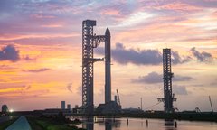 The image is a highly blurred, vertically stretched photo showing silhouettes of tall tower-like structures (likely industrial or launch-related towers) against a dramatic sunset sky with pink, orange, and purple clouds reflected in water below. The caption: "A blurred sunset scene with silhouetted towers looms over reflective waters, mirroring the uncertain and painful horizon for SpaceX bulls whose leveraged long positions on Hyperliquid are now deeply in the red following the company's drop below its IPO price."