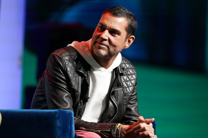 A man with a beard wearing a black quilted leather jacket and white hoodie sitting on a blue chair.