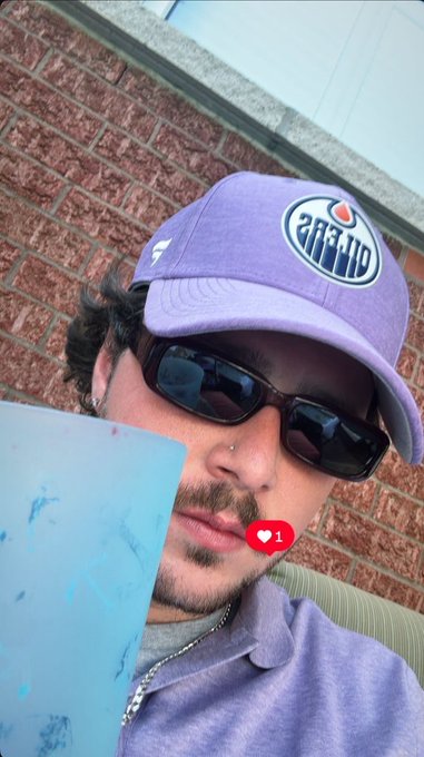A young man wearing a purple "KINGS" NBA cap, black sunglasses, and a small heart "1" reaction icon poses for a selfie while sipping from a blue cup against a brick wall backdrop, channeling a casual Friday vibe to match the "GM happy Friday" greeting.