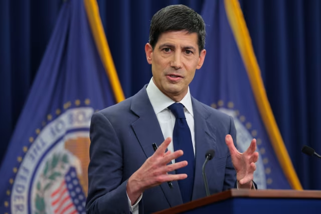 A man in a blue suit and tie speaks at a podium, gesturing with both hands, set against a backdrop featuring flags and the Federal Reserve seal. The image complements the post's discussion of the Federal Reserve's "hawkish hold" and its impact on Bitcoin and broader markets, visually representing the central figure in monetary policy decisions influencing crypto sentiment.