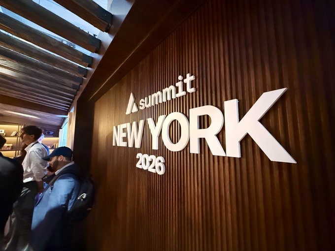 A photo taken at an event showing a wooden wall with large white dimensional lettering that reads 'summit NEW YORK 2026', with attendees standing to the left.