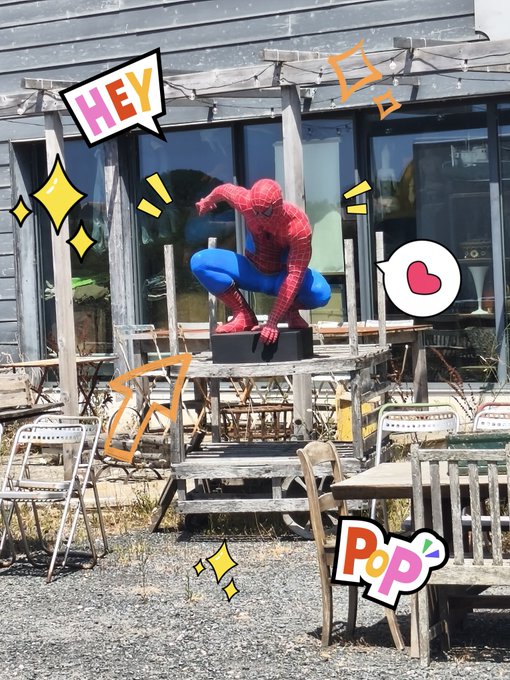 A person in a Spider-Man costume crouching on a wooden table outdoors, with playful comic-style stickers including 'HEY', 'Pop', a heart, stars, and action lines decorating the photo.