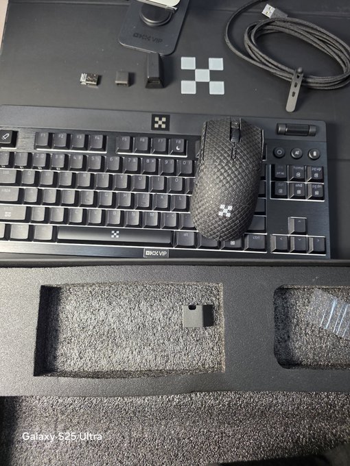 A photo of unboxed gaming peripherals including a black keyboard, mouse, USB receiver, and braided cable laid out in foam packaging, taken with a Galaxy S25 Ultra.